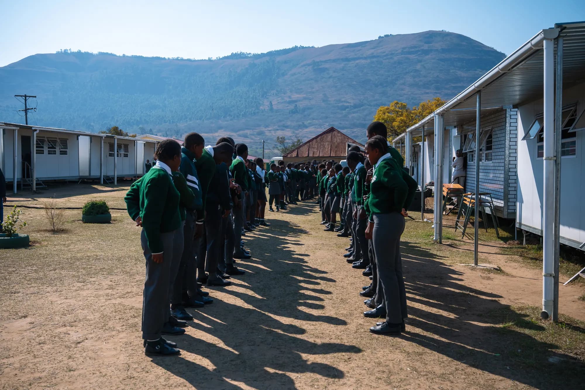 Learners standing in assembly lines between classroom blocks, hills rising behind
